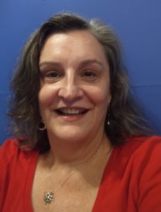 A woman with greying hair and a red shirt, background is a blue wall. 